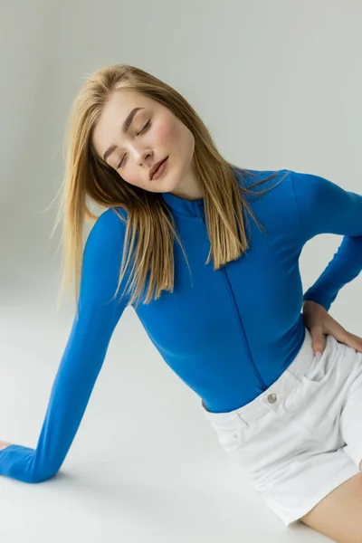blonde woman in blue zipped turtleneck sitting with closed eyes and hand on hip on grey background