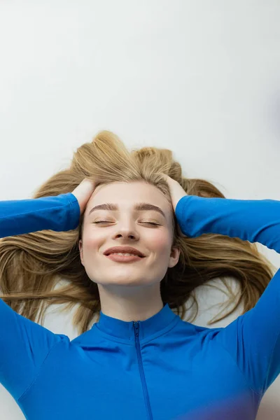 top view of young blonde woman smiling with closed eyes while lying on grey background