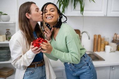 happy lesbian woman kissing cheek of pleased african american girlfriend holding red gift box on valentines day clipart