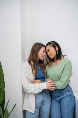 Evdeki yeşil bitkinin yanında sarılıp birbirlerine bakan ırklar arası ve lezbiyen kadınlar.