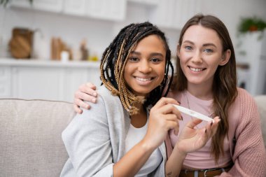 tattooed african american and lesbian woman holding pregnancy test near cheerful girlfriend  clipart