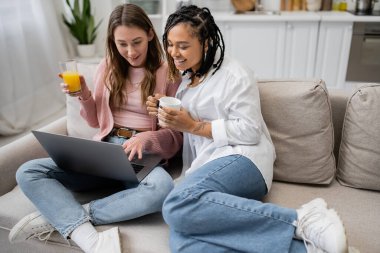 happy african american lesbian woman holding cup of coffee near girlfriend using laptop while working from home  clipart