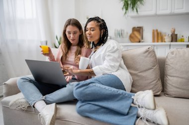 Afro-Amerikan lezbiyen kadın, kız arkadaşının yanında elinde kahve fincanı tutarken evde çalışırken dizüstü bilgisayar kullanıyor. 