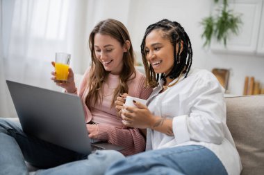 young african american lesbian woman holding cup of coffee near happy girlfriend using laptop while working from home  clipart