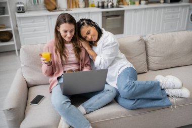 young african american lesbian woman leaning on girlfriend using laptop while working from home  clipart