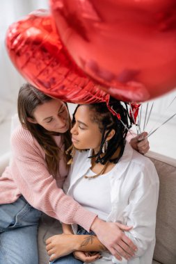 overhead view of lesbian woman holding red balloons and sitting on couch with tattooed african american girlfriend clipart