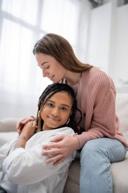 Mutlu ve lezbiyen kadın kanepede oturuyor ve dövmeli Afro-Amerikan kız arkadaşını kucaklıyor.