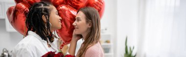interracial lesbian women looking at each other near red roses and balloons on valentines day, banner  clipart