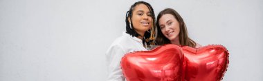 Kırmızı kalp şeklinde balonlar taşıyan neşeli ve ırklar arası lezbiyen kadınlar gri pankartta izole edilmiş.