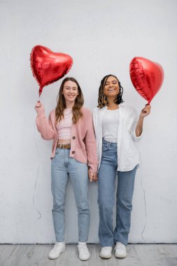 Gri üzerinde kırmızı kalp şeklinde balonlarla yüzleri kaplayan mutlu ırklar arası lezbiyen kadınlar. 