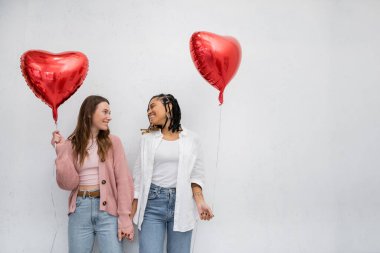 cheerful and interracial lesbian women holding red heart-shaped balloons and looking at each other isolated on grey  clipart