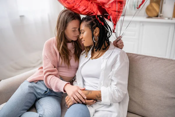 sensual lesbian woman holding red balloons and sitting on couch with african american girlfriend on valentines day