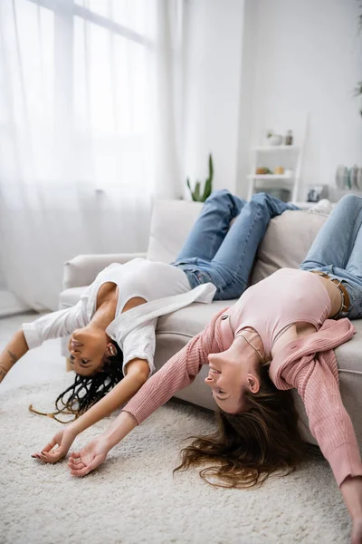 positive and multiethnic lesbian couple lying upside down on couch in living room