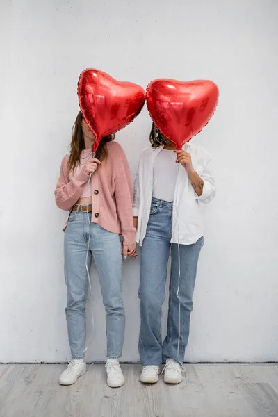 stock image interracial lesbian women covering faces with red heart-shaped balloons on grey 