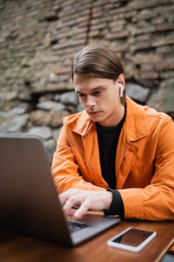 Kulaklıklı genç serbest çalışan açık kafede akıllı telefonun yanında dizüstü bilgisayar kullanıyor. 