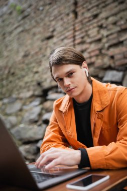 Kulaklık takan, dizüstü bilgisayar kullanan ve dışarıdaki kafede kameraya bakan genç bir serbest yazar. 