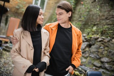 Genç adam açık kafede trençkotlu gülümseyen Asyalı kız arkadaşına sarılıyor. 