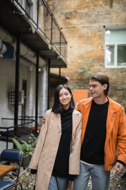 Asyalı kız arkadaşına sarılan şık bir genç adam. Açık kafede trençkot giymiş. 