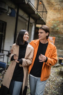 Havalı bir adam açık kafede neşeli Asyalı kız arkadaşıyla sarılıp konuşuyor. 