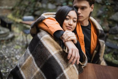 Genç adam kafenin terasında battaniyeli Asyalı kız arkadaşına sarılıyor. 