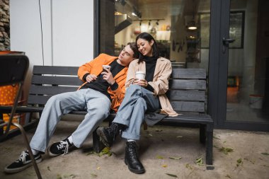 Kahve tutan şık bir adam, kafenin terasındaki bankta oturan neşeli Asyalı kız arkadaşının yanına gitmek için. 