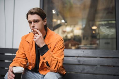Tarz sahibi bir adam sigara içiyor ve kafenin yakınına gitmek için kahve tutuyor. 