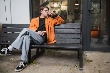 Stylish man smoking cigarette and holding coffee to go near cafe  clipart