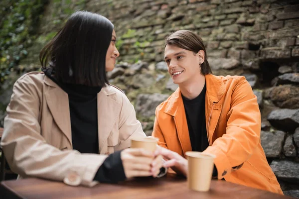 Açık kafede elinde kağıt bardakla Asyalı kız arkadaşının eline dokunan gülümseyen adam. 