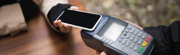 Cropped view of customer paying with mobile phone near waiter with payment terminal in cafe, banner 