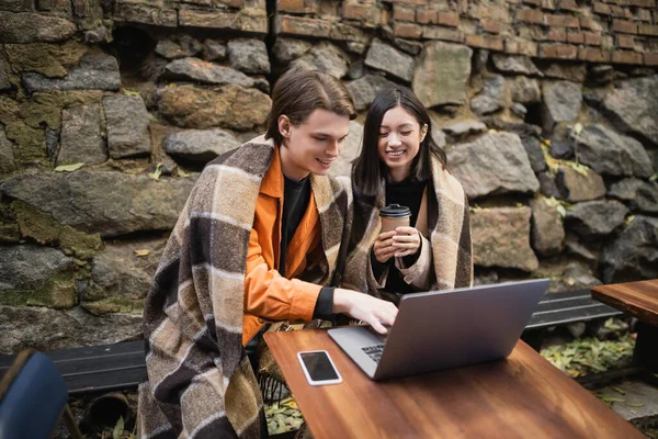 Kafenin terasında dizüstü bilgisayar kullanan çok ırklı bir çift var. 