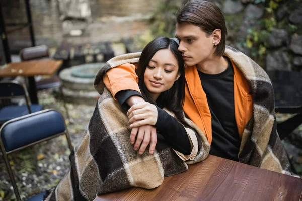 Genç adam kafenin terasında battaniyeli Asyalı kız arkadaşına sarılıyor. 