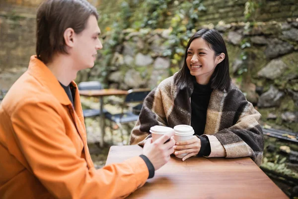 Smiling asian woman in blanket holding coffee to go and looking at boyfriend in outdoor cafe