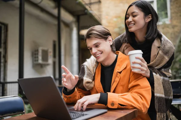 Kahvesini elinde tutan neşeli Asyalı kadın, erkek arkadaşına sarılmak için terasta dizüstü bilgisayar kullanıyor. 
