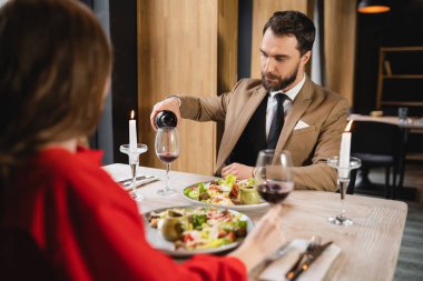 bearded man pouring red wine in glass near girlfriend during dinner on valentines day clipart