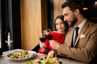 young woman and cheerful man clinking glasses with red wine in restaurant on valentines day  clipart