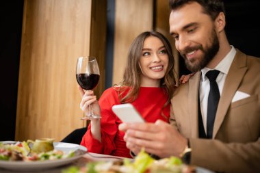 cheerful man holding smartphone near happy girlfriend with glass of wine during celebration on valentines day  clipart