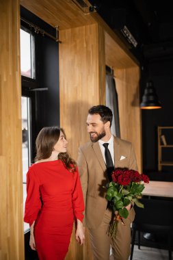 bearded man holding red roses while looking at cheerful girlfriend in dress on valentines day clipart