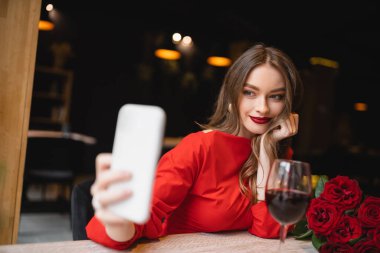 Sevgililer gününde şarap ve kırmızı güllerin yanında selfie çeken neşeli genç bir kadın.