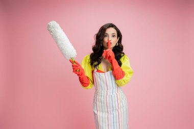 brunette woman in yellow blouse and red rubber gloves holding white feather duster and showing hush sign isolated on pink clipart