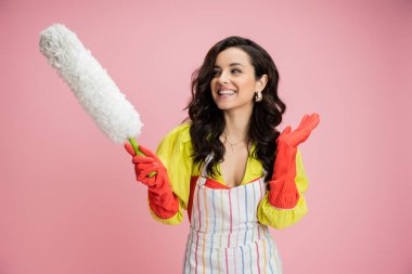 Çizgili önlüklü gülümseyen ev hanımı ve beyaz toz fırçası tutan kırmızı lastik eldivenler ve pembe izole edilmiş el sallayan el sallayan.