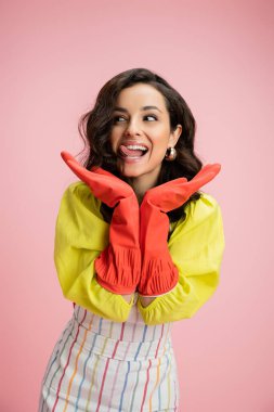 excited housewife in yellow blouse and red rubber gloves holding hands near face and sticking out tongue isolated on pink clipart