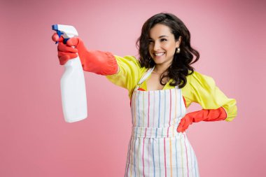 smiling brunette housewife standing with hand on hip and spraying from bottle isolated on pink clipart