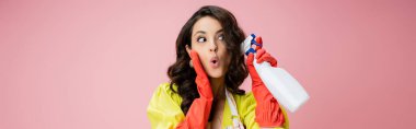 thrilled brunette woman in red rubber gloves holding spray bottle and looking away isolated on pink, banner clipart