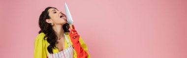 pretty brunette housewife in yellow blouse and red rubber glove licking kitchen knife isolated on pink, banner clipart