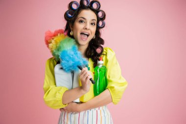 astonished housewife in hair curlers holding detergents and bright feather duster isolated on pink clipart