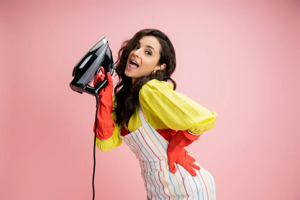 stock image amazed and flirty housewife in yellow blouse and apron holding iron and looking at camera isolated on pink