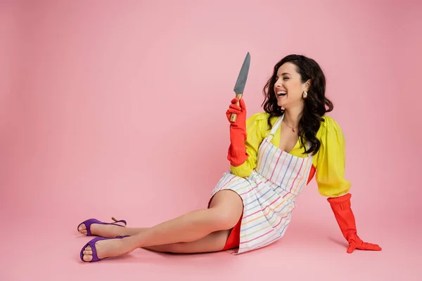 stock image happy housewife in striped apron and sandals holding kitchen knife while sitting on pink background