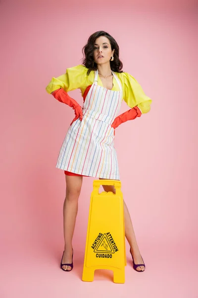 full length of confident and stylish housewife standing with hands on hips near wet floor sign on pink background