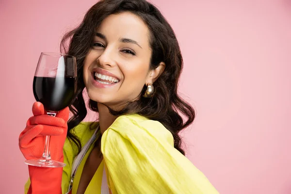 stock image portrait of happy housewife with brunette wavy hair wearing red rubber glove and holding wine glass isolated on pink