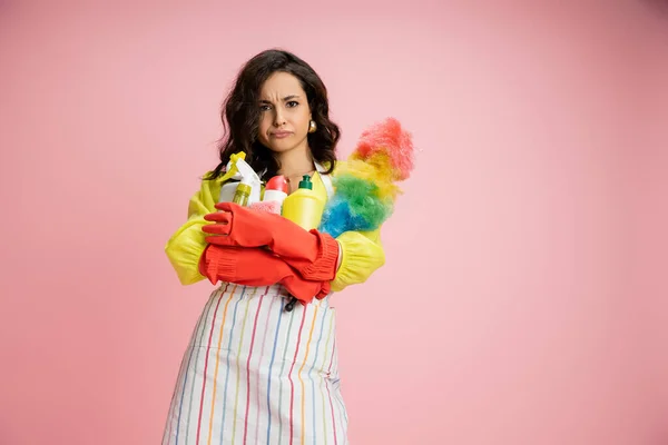 stock image frowning housewife in striped apron and red rubber gloves holding pile of cleaning supplies isolated on pink